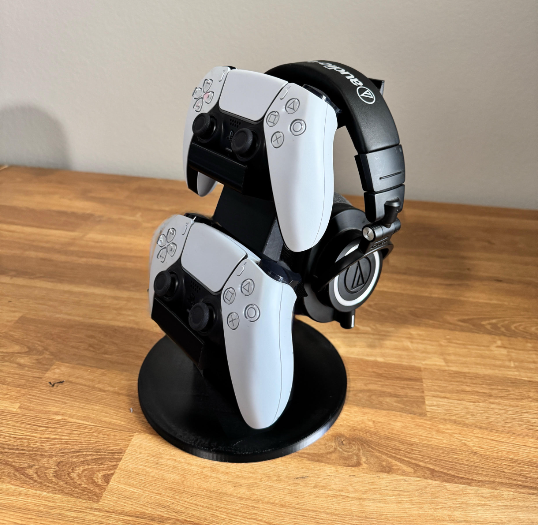 Black 3D-printed stand holding two white DualSense controllers and over-ear headphones on a wooden desk